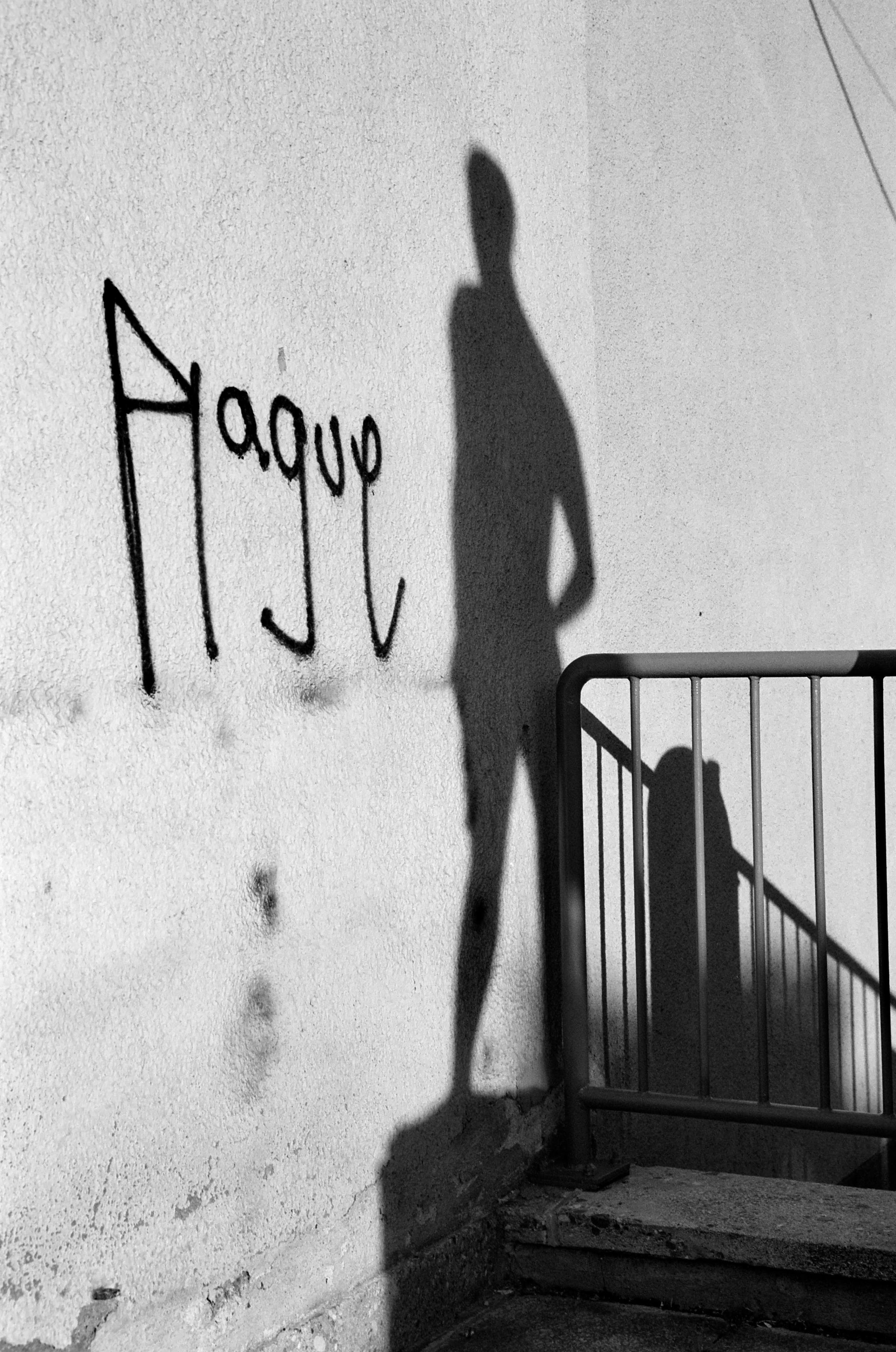The black and white image shows the shadow of a standing person cast on a wall beside graffiti, next to a stair railing.