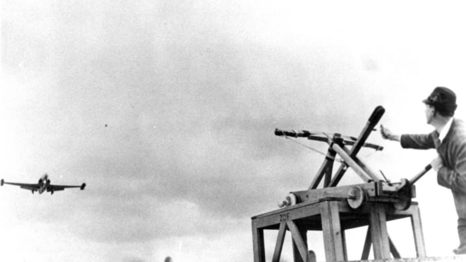 Schwarz-Weiß-Foto: 
Der Knödelschütz visiert mit seinem Katapult ein im Tiefflug herannahendes Flugzeug an, das nach Expertenmeinung aber kein Starfighter, sondern eine Fouga Magister ist. 