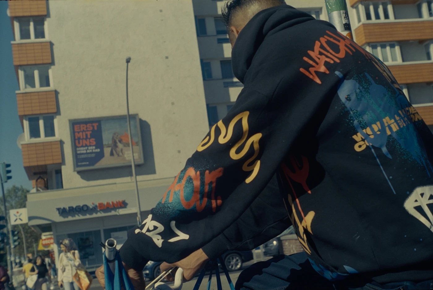 eine person auf einem fahrrad mit einem auffällig schwarzbunten hoodie steht an einer ampel, stark angeschnitten von unten fotografiert, sodass nur oberkörper und hinterkopf zu sehen ist. im hintergrund stadtlandschaft.