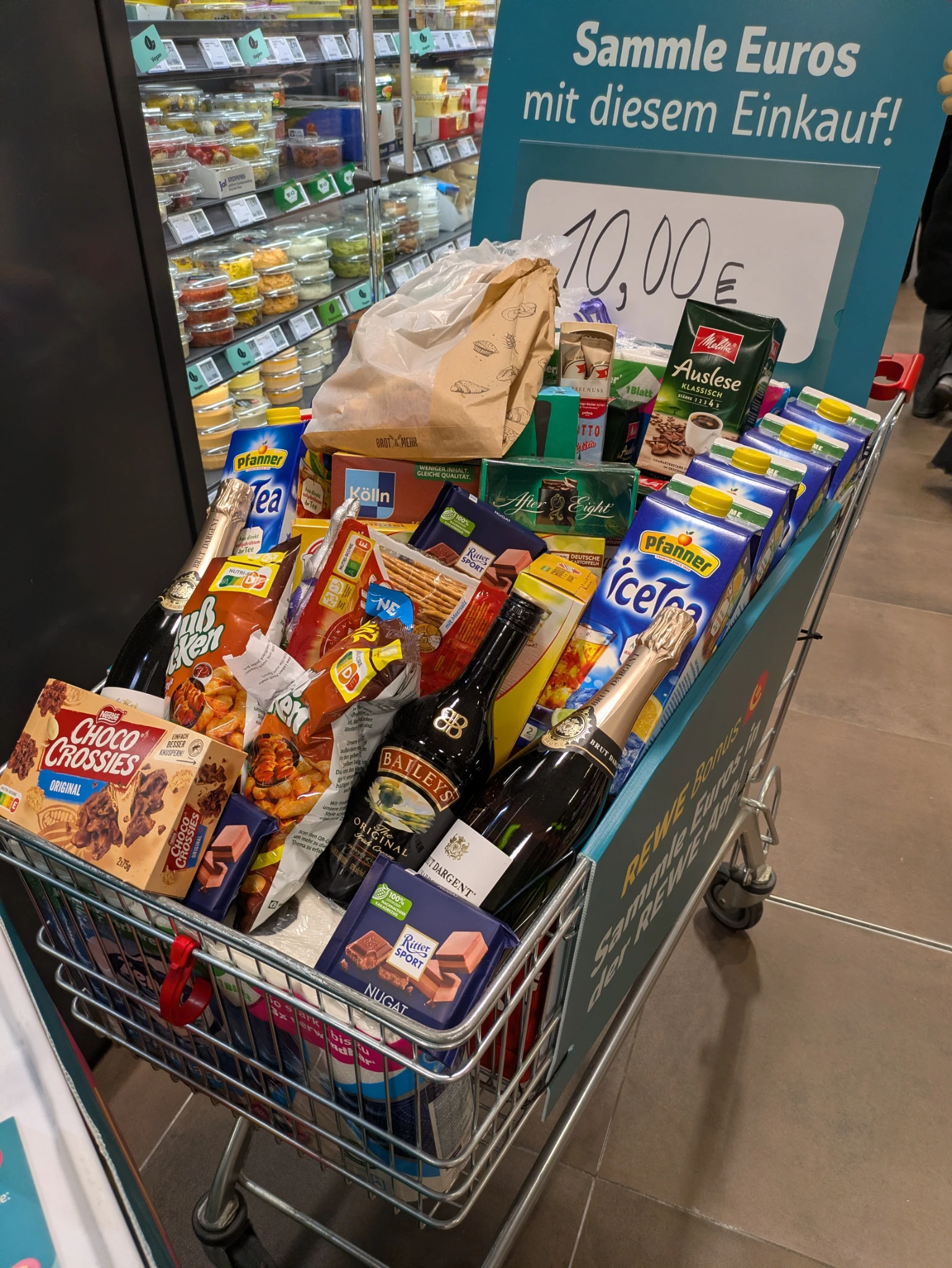 ein gefüllter einkauswagen in einem supermarkt, dahinter ein schild:

Sammle Euros mit diesem Einkauf 
10,00 €

der korb iat voll eistee, alkohol und süßigkeiten