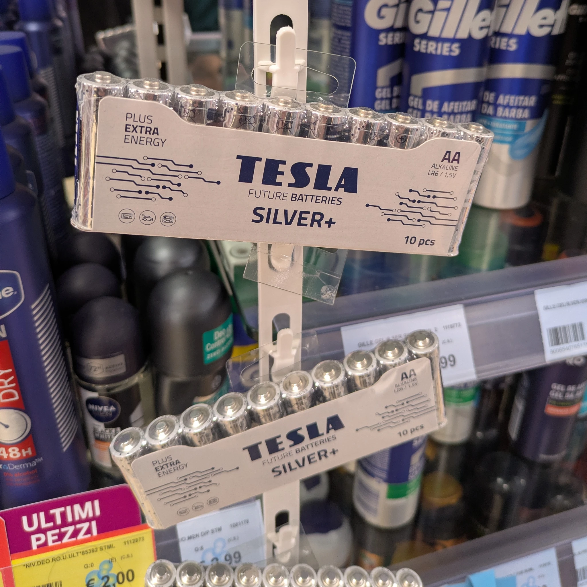 an einem regal im supermarkt hängen zwei packungen AA-Batterien der Marke tesla. im Hintergrund andere produkte
