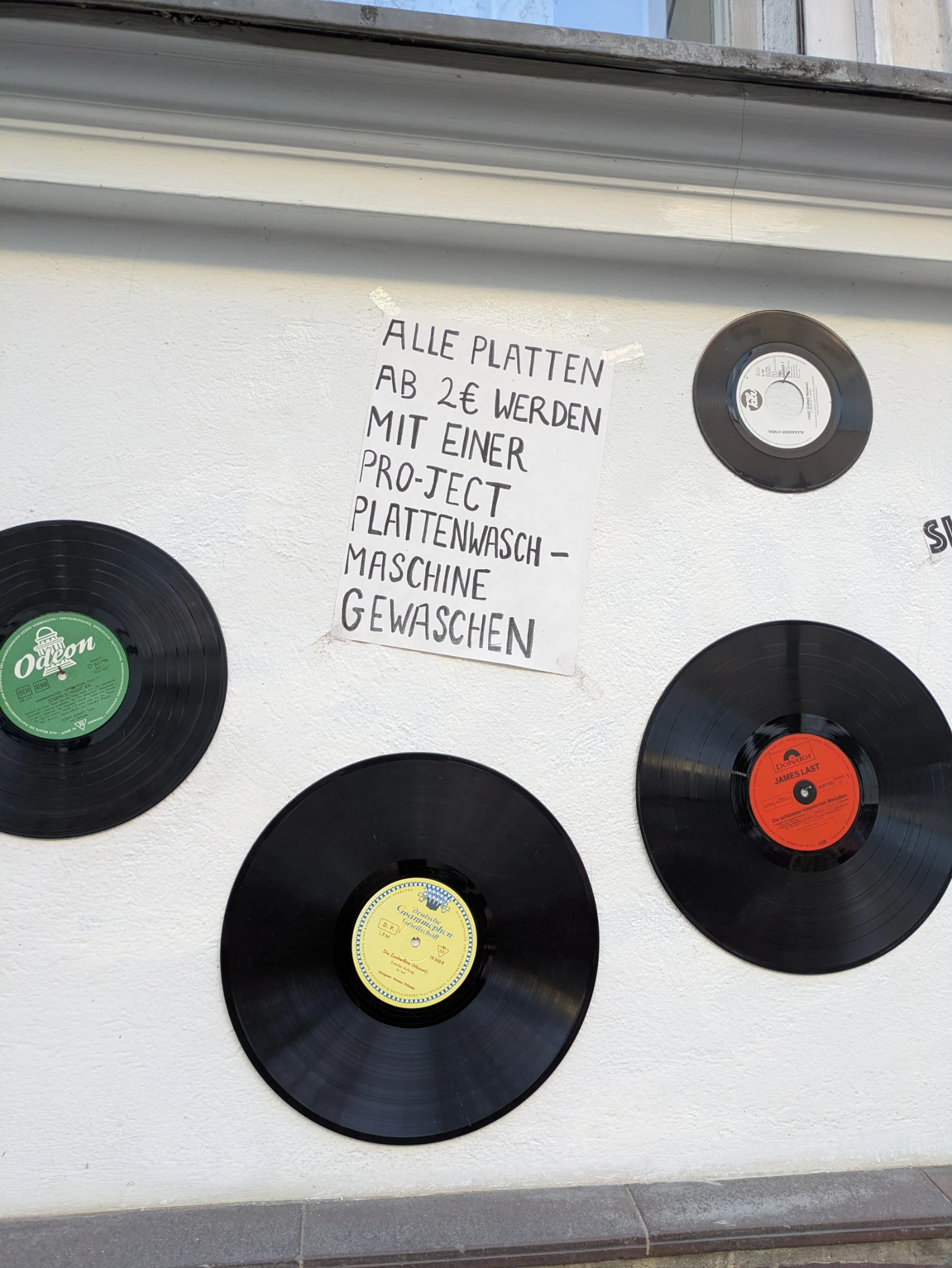 auf einer hauswand drei schallplatten und ein handgeschriebenes schild:

ALLE PLATTEN
AB 2€ WERDEN
MIT EINER
PRO-JECT
PLATTENWASCH-
MASCHINE
GEWASCHEN