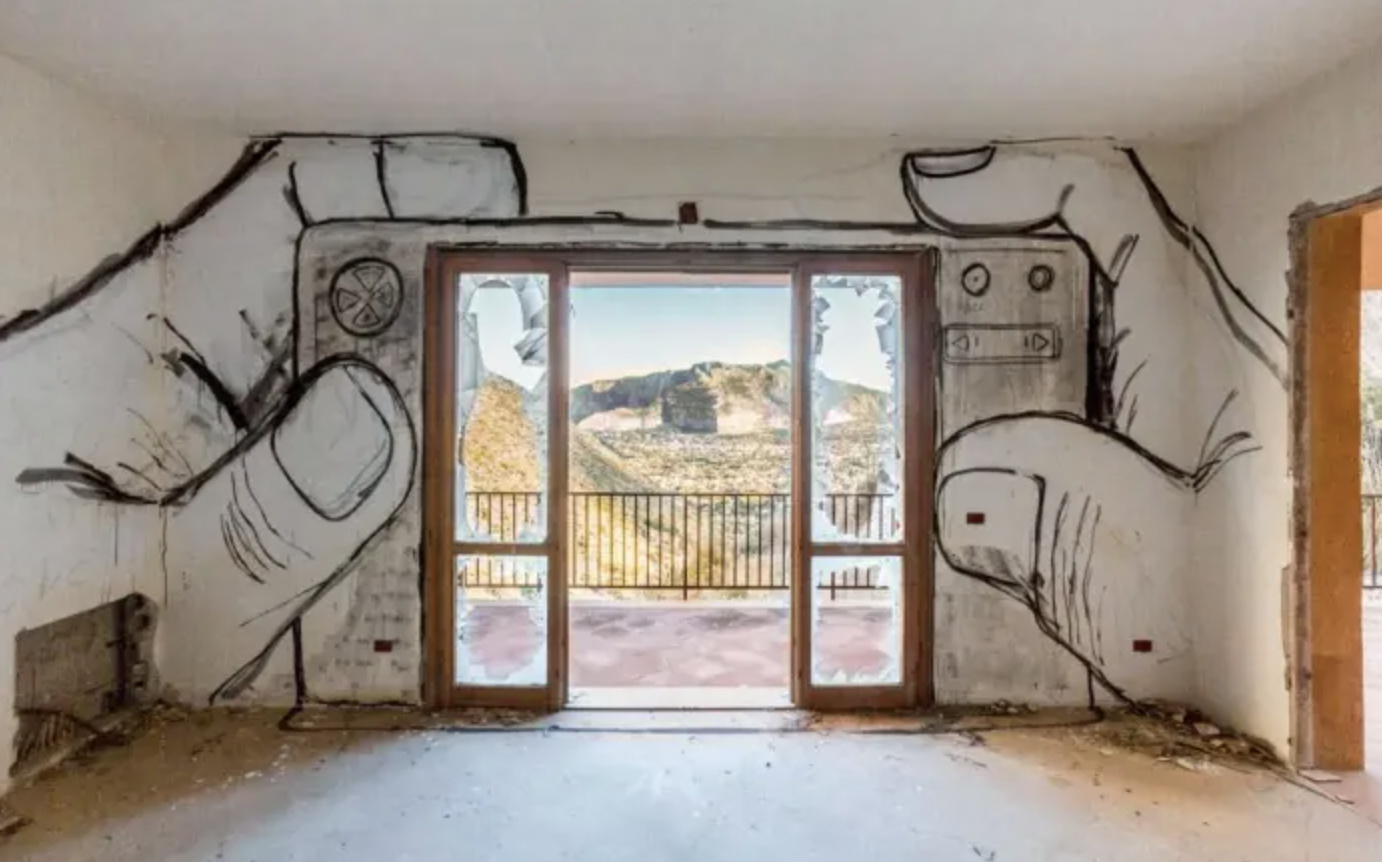 A black and white mural of two hands holding a camera around a balcony doorway. Painted by Collettivo FX at the Pizzo Sella Art Village in Palermo, Italy. The window acts as a natural lens framing the mountains.