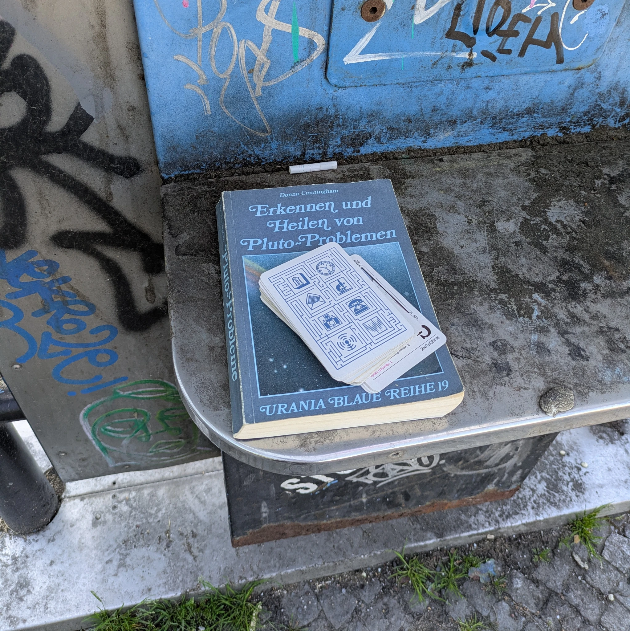 in einer alten und ehemaligen telefonzelle (man sieht nur einen teil davon) liegt ein buch mit dem titel

Erkennen und Heilen von Pluto-Problemen

darauf ein stapel spielkarten