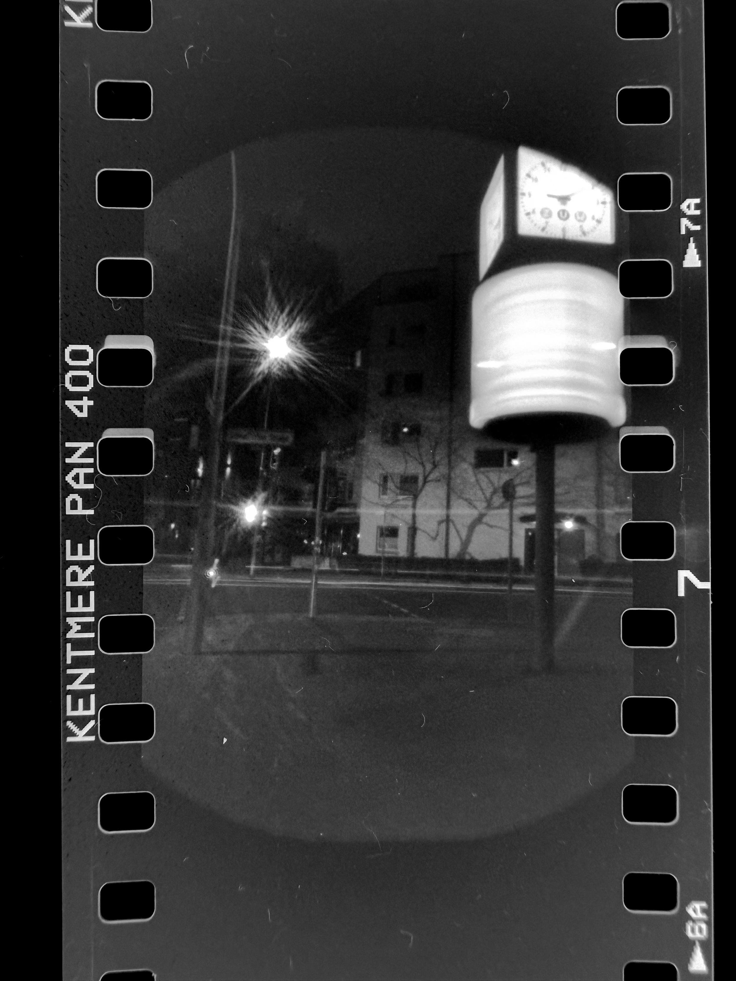 Ein Scan von einem Film. Darauf zu sehen eine Nachtaufnahme von einer Uhr im öffentlichen Raum mit drehender Werbung drauf die bewegungsunscharf geworden ist. 