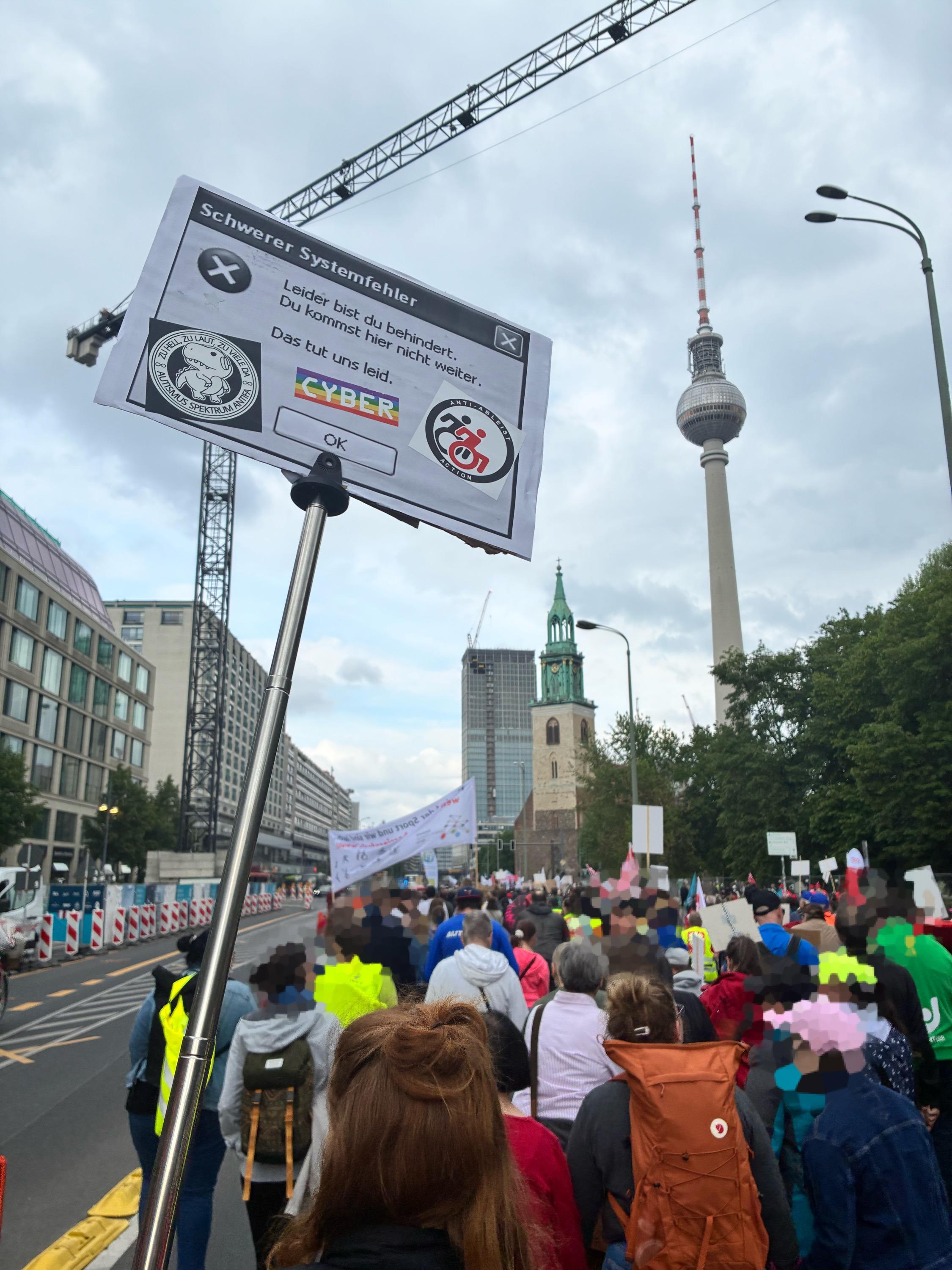 Foto eines langen Demo-Zuges entlang „Unter den Linden“ in Berlin, mit dem Fernsehturm im Blick. Der Himmel ist bewölkt und mein Demoschild ragt über die Menschen. 

Mein Demoschild: Windows XP Fehlerneldung mit dem Titel: „Schwerer Systemfehler“ und dem Text „Leider bist du behindert. Du kommst hier nicht weiter. Das tut uns leid.“. Zum Bestätigen gibt es nur „OK“.

Darauf kleben „Anti Ableist Action“ mit Rollstuhl-Symbolen im Antifa-Design, dem Sticker „zu hell, zu laut, zu viele da, Autismus Spektrum antifa“ mit einem Dino, und einem regenbogenfarbenen Cyber-Sticker.