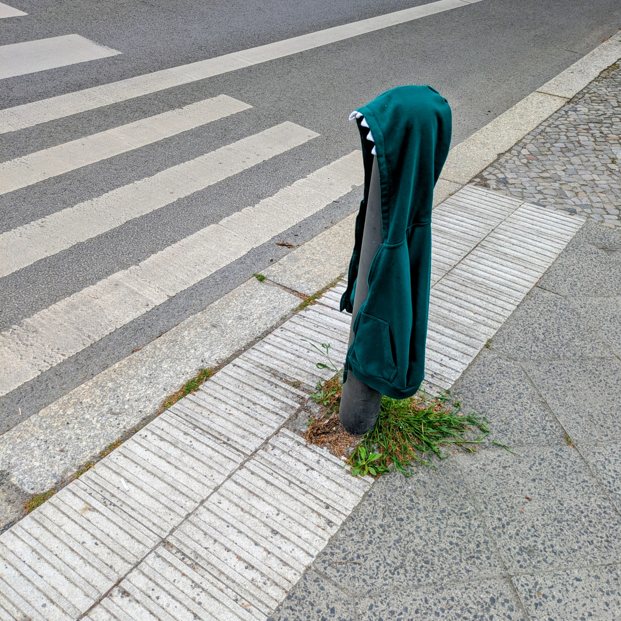 ein stück straße mit zebrastreifen, ein stück fußweg, ein poller. 

auf dem poller hängt ein grüner hoodie für kinder. an der kapuze vorn weiße zähne.