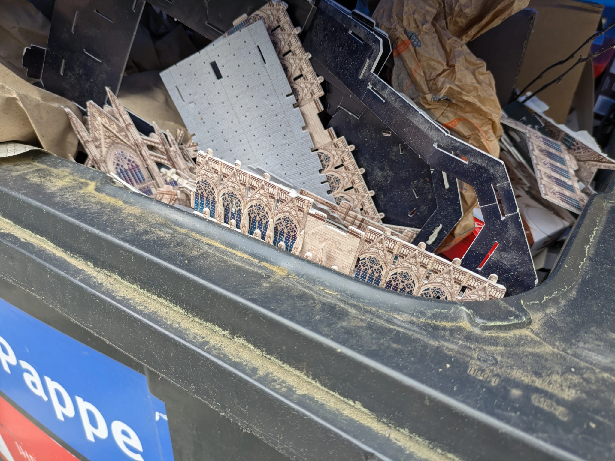 nahaufnahme eines halb geöffneten papiercontainers, es wurden teile eines pappbausatzes (3d puzzle?) weggeworfen, sie sehen aus wie die kirche notre dame in paris
