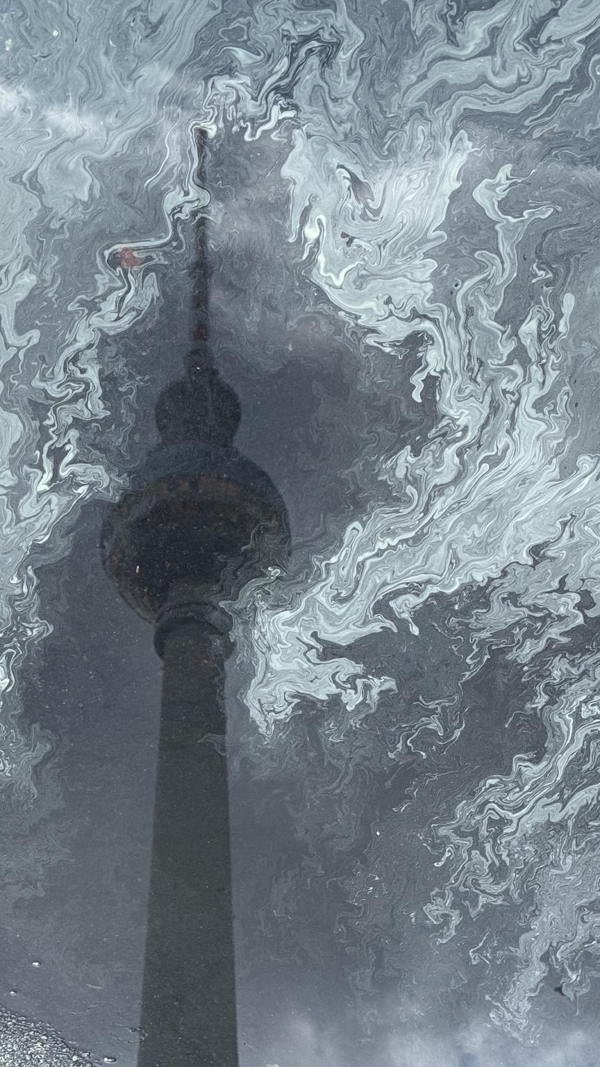 Reflection of a tower amidst swirling patterns on a water surface, with cloudy skies in the background.