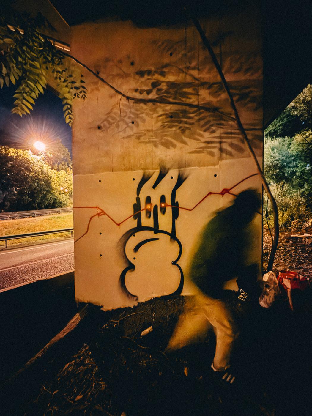 Graffiti of a cartoonish face on a concrete wall under a bridge at night, with a blurred figure walking past and trees in the background.