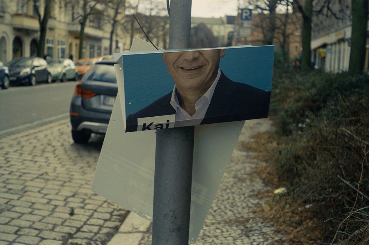 ein wahlplakat im urbanen raum. das plakat ist zusammen geknickt, wir sehen nur ein teil des regierenden bürgermeister berlins kai wegner, darunter steht Kai. im hintergrund eine straße mit kopfsteinpflaster und parkende autos.