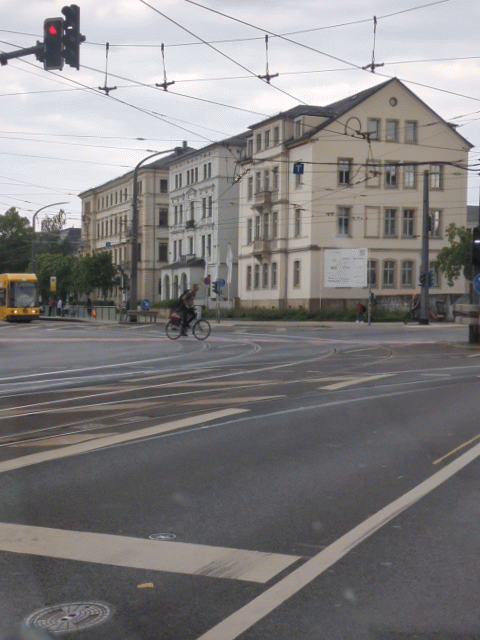 ein animiertes gif, eine Straßenkreuzung im urbanen raum, typ mit riesigem plüschbär auf dem rücken fährt mit fahrrad quer über die kreuzung