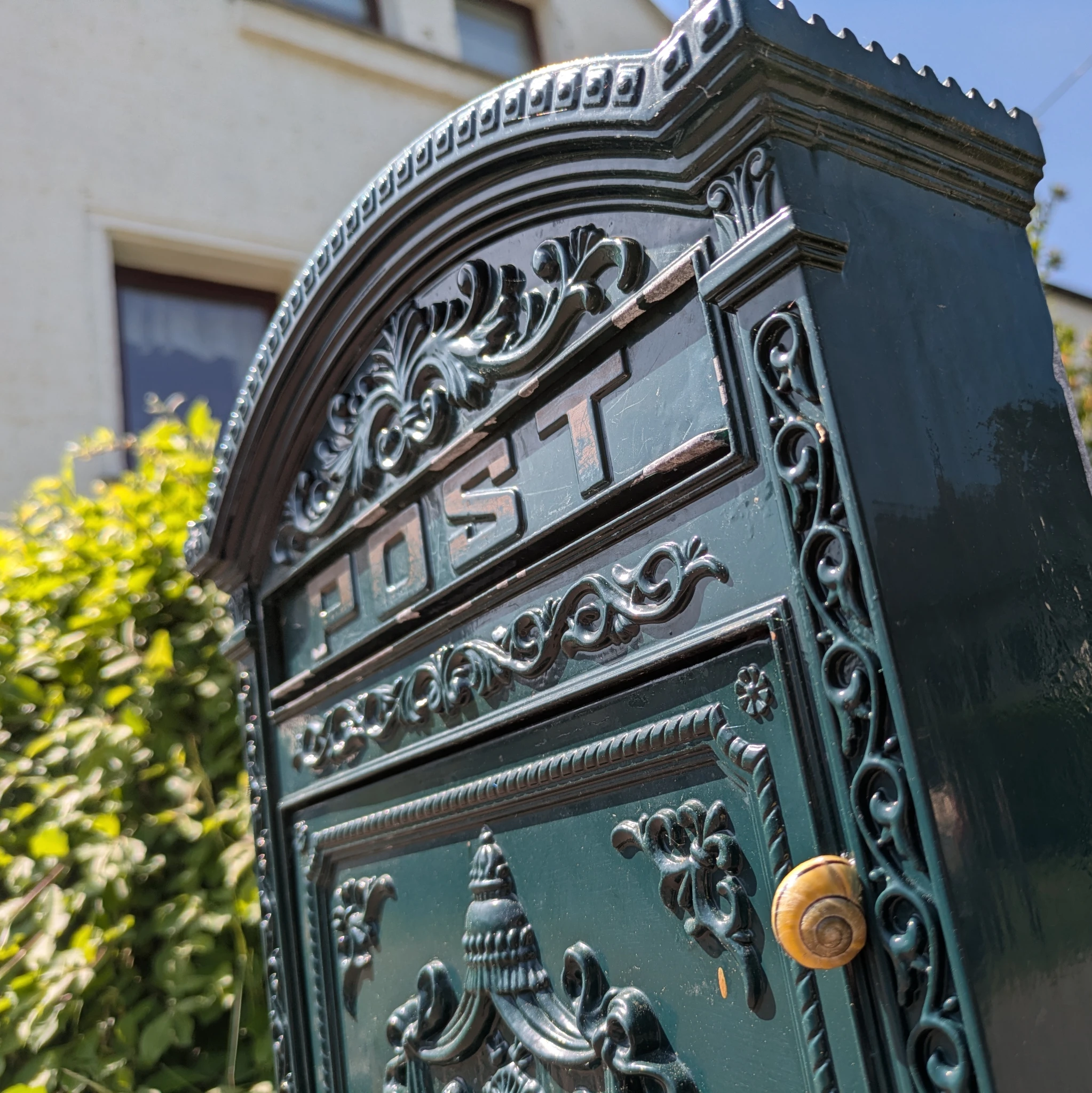 eine schnecke hängt an einem reich verzierten briefkasten, auf dem POST steht. im Hintergrund ein haus und pflanzen.