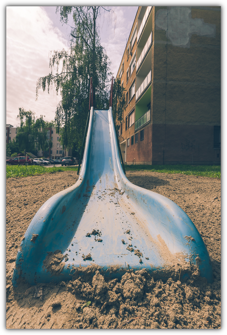 Eine blaue Rutsche auf sandigem Boden, dahinter mehrere Wohngebäude undgroße Bäume unter leicht bewölktem Himmel.