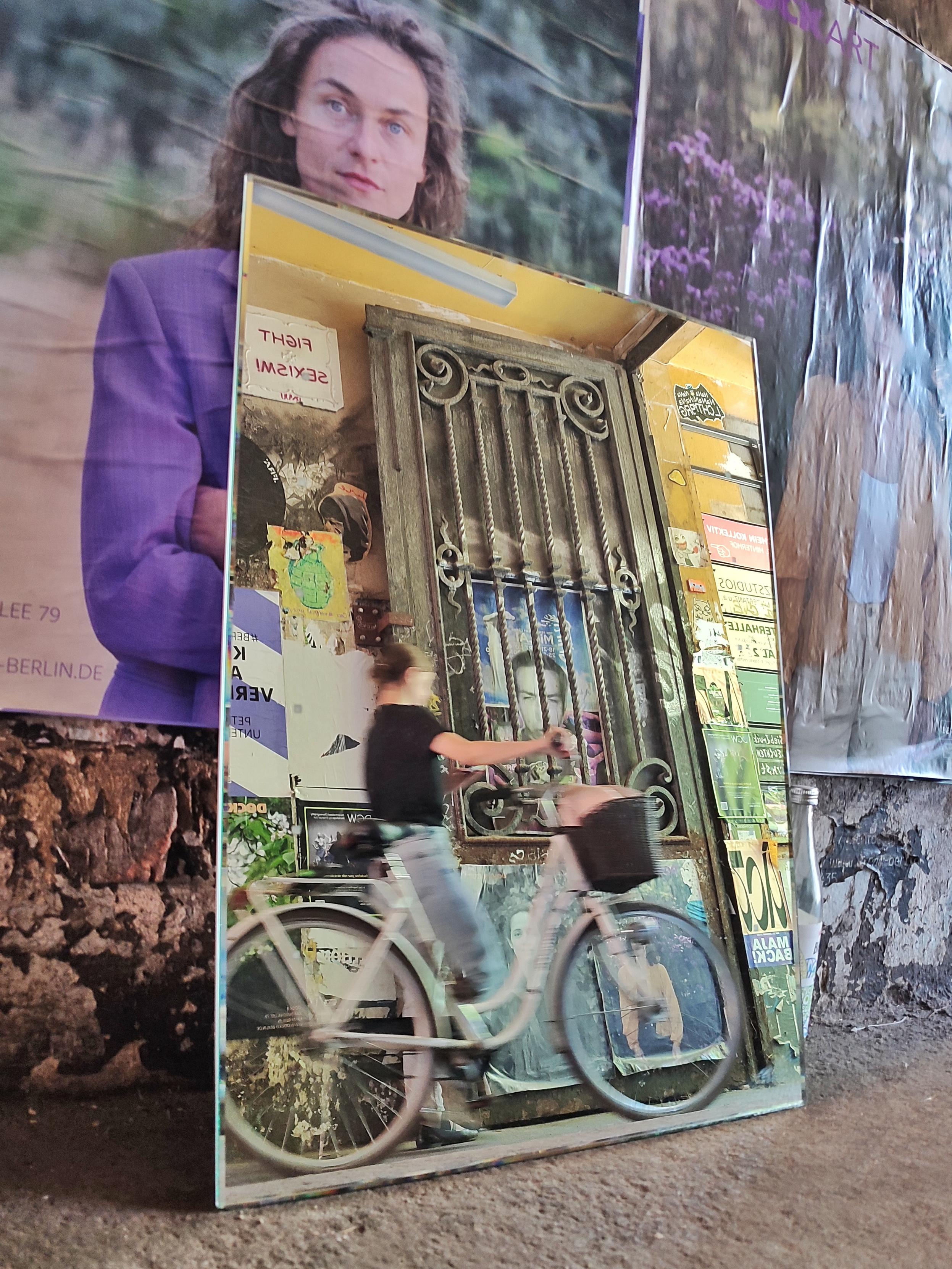 Farbfoto im Hochformat. An einer mit Plakaten bedecken Wand lehnt ein großer Spiegel, darin ist schemenhaft eine vorbeigehende Frau mit einem Fahrrad zu erkennen, außerdem die gegenüberliegende Wand mit Graffiti und einem Metalltor. Hinter dem Metalltor klebt ein Poster an der Wand, es zeigt einen Männerkopf. Auf dem Poster hinter dem Spiegel ist eine Frau abgebildet, sie schaut direkt in die Kamera.