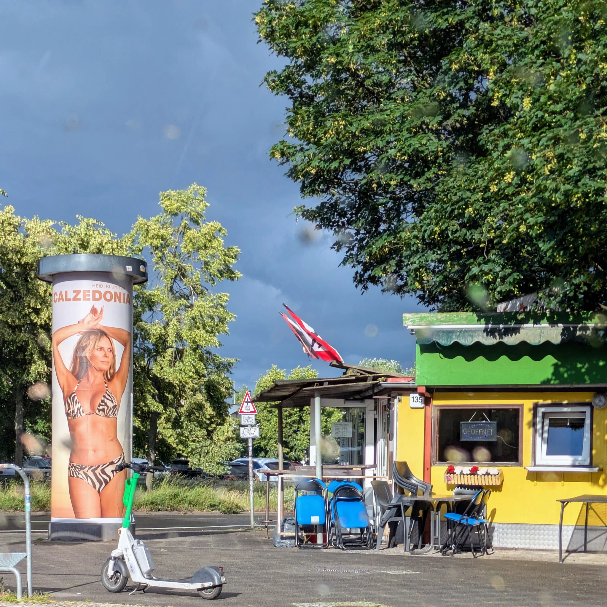eine straßenszene, ein gelb grüner kiosk/imbiss mit gestapelten stühlen davor, darüber thront ein grüner baum und links eine litfaßsäule mit heidi klum im bikini.