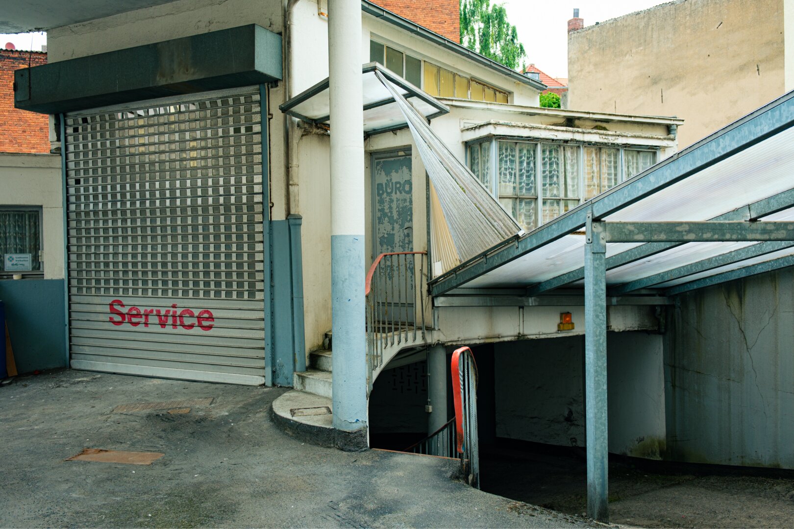An urban scene showing a service entrance with a metal rolling shutter partially closed. There is a stairway leading down and a covered walkway with a slanted roof. The surrounding walls are weathered, and the overall atmosphere is somewhat neglected.