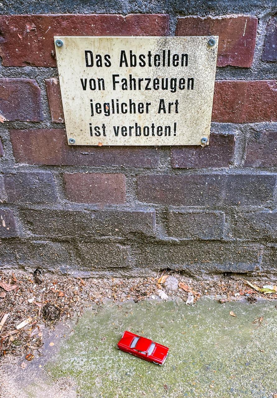 Foto vom unteren Bereich eines gemauerten Wohnhauses sowie dem davor befindlichen Gehweg. Ungefähr in Wadenhöhe ist ungewöhnlich tief ein Schild angebracht, auf dem steht: das Abstellen von Fahrzeugen jeglicher Art ist verboten! Direkt unter dem Schild auf dem Boden steht ein kleines rotes Matchbox-Auto (amerikanischer Straßenkreuzer aus den späten 1950er-Jahren, den hab natürlich ich extra für das Foto dahingestellt).