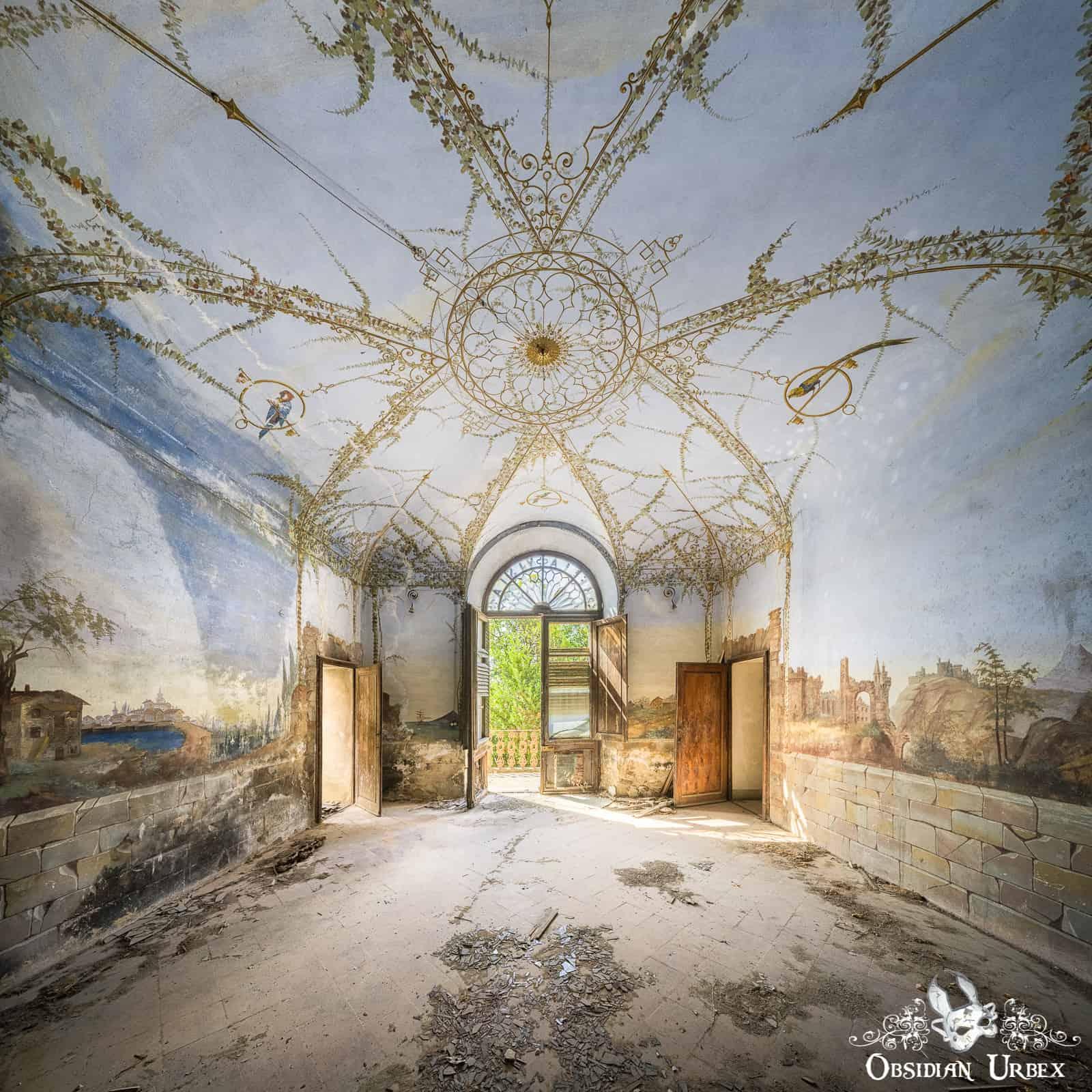 An abandoned Italian villa's entrance hall features a vaulted ceiling with intricate golden vine frescoes and wall murals of landscapes. An open doorway leads to the outside.