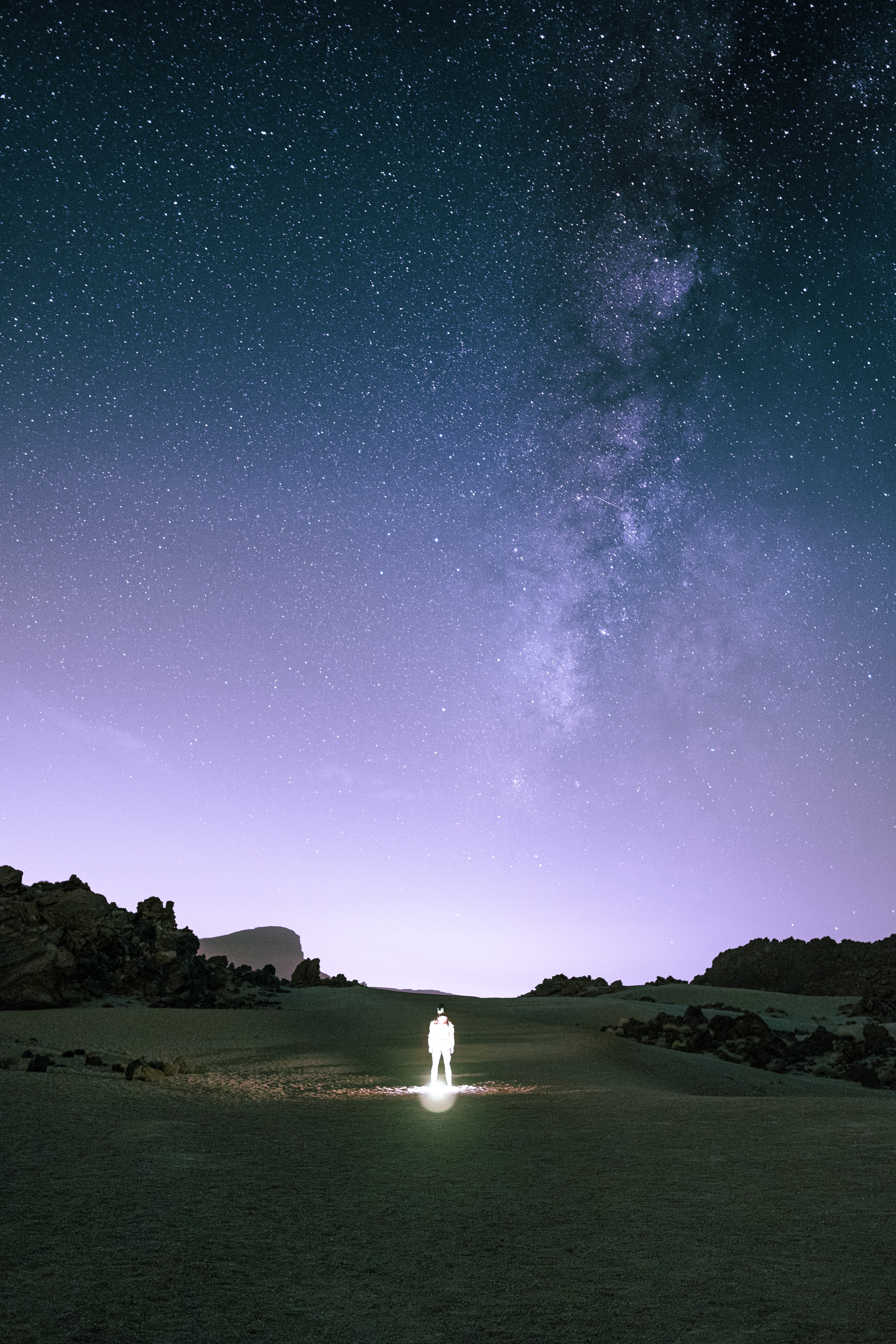 A person illuminated by a headlamp standing alone in a dark volcanic valley on Tenerife, Spain, dwarfed beneath a vast starry sky with the Milky Way stretching overhead.