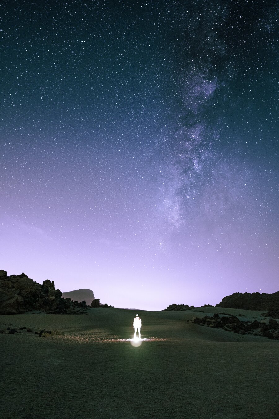 A person illuminated by a headlamp standing alone in a dark volcanic valley on Tenerife, Spain, dwarfed beneath a vast starry sky with the Milky Way stretching overhead.