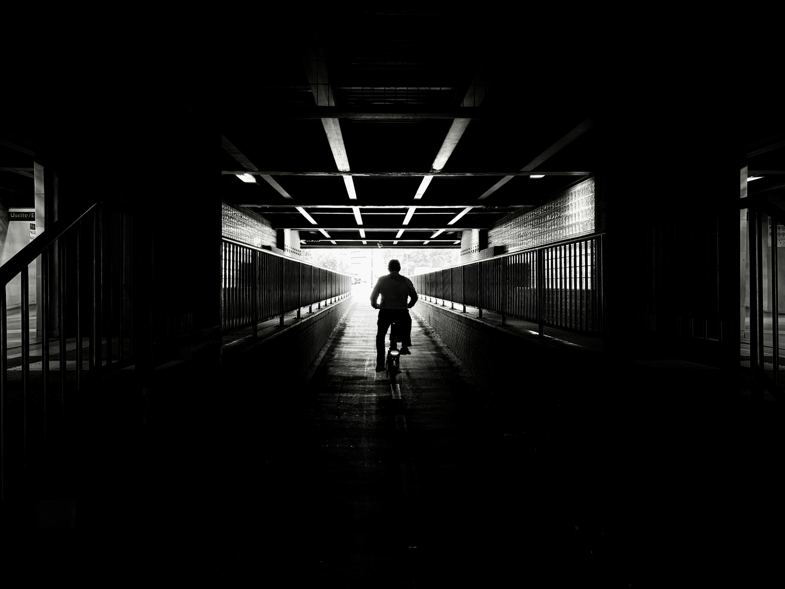 Black and white photo of a person riding a bicycle through the Balduina underpass. The cyclist is silhouetted against the bright light at the tunnel's exit. The underpass has a low ceiling with exposed beams and metal railings lining both sides of the path. The foreground is dark, emphasizing the contrast with the light at the end of the tunnel.