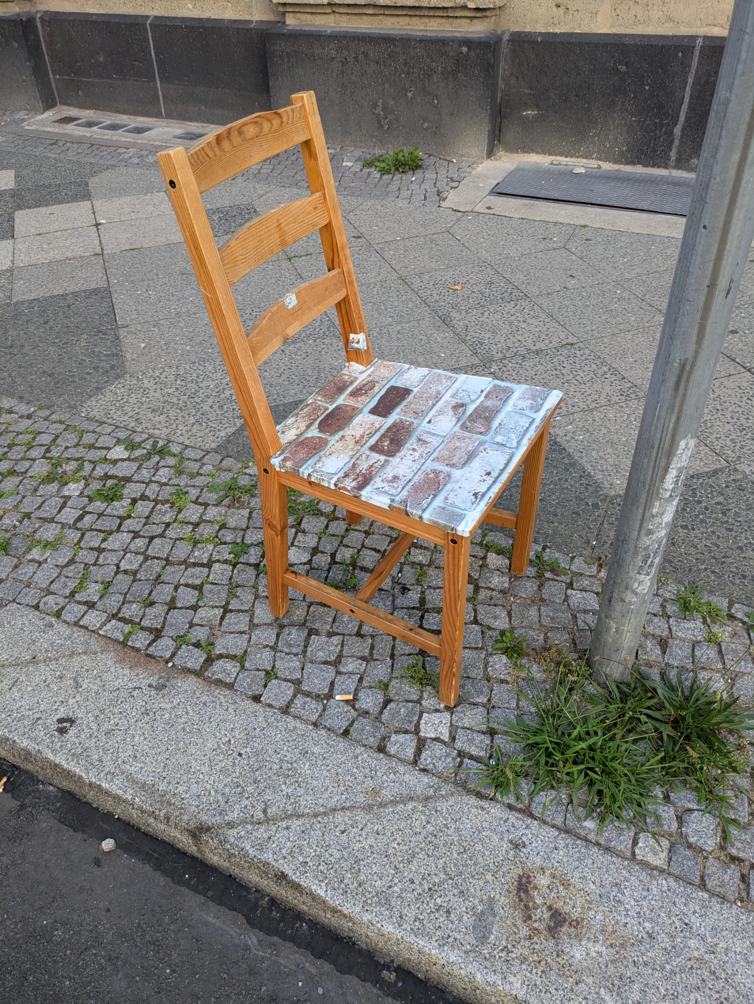 ein einsamer küchenstuhl aus holz steht auf einem bürgersteig. die sitzfläche ist mit einer folie beklebt, die eine steinmauer zeigt.