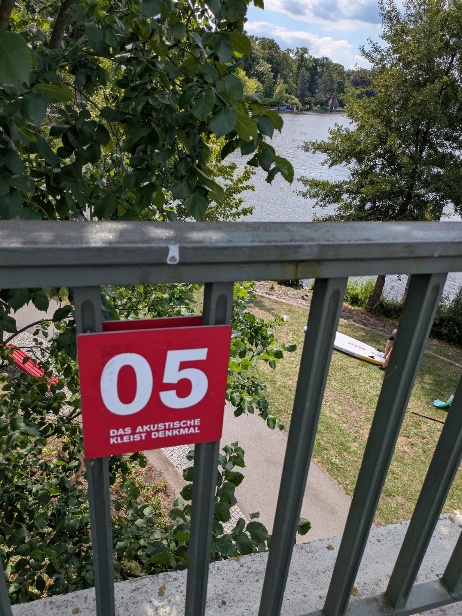 blick von einer brücke auf viel grün und wasser, am Brückengeländer ein rotes schild 

05
Das akustische Kleist-Denkmal