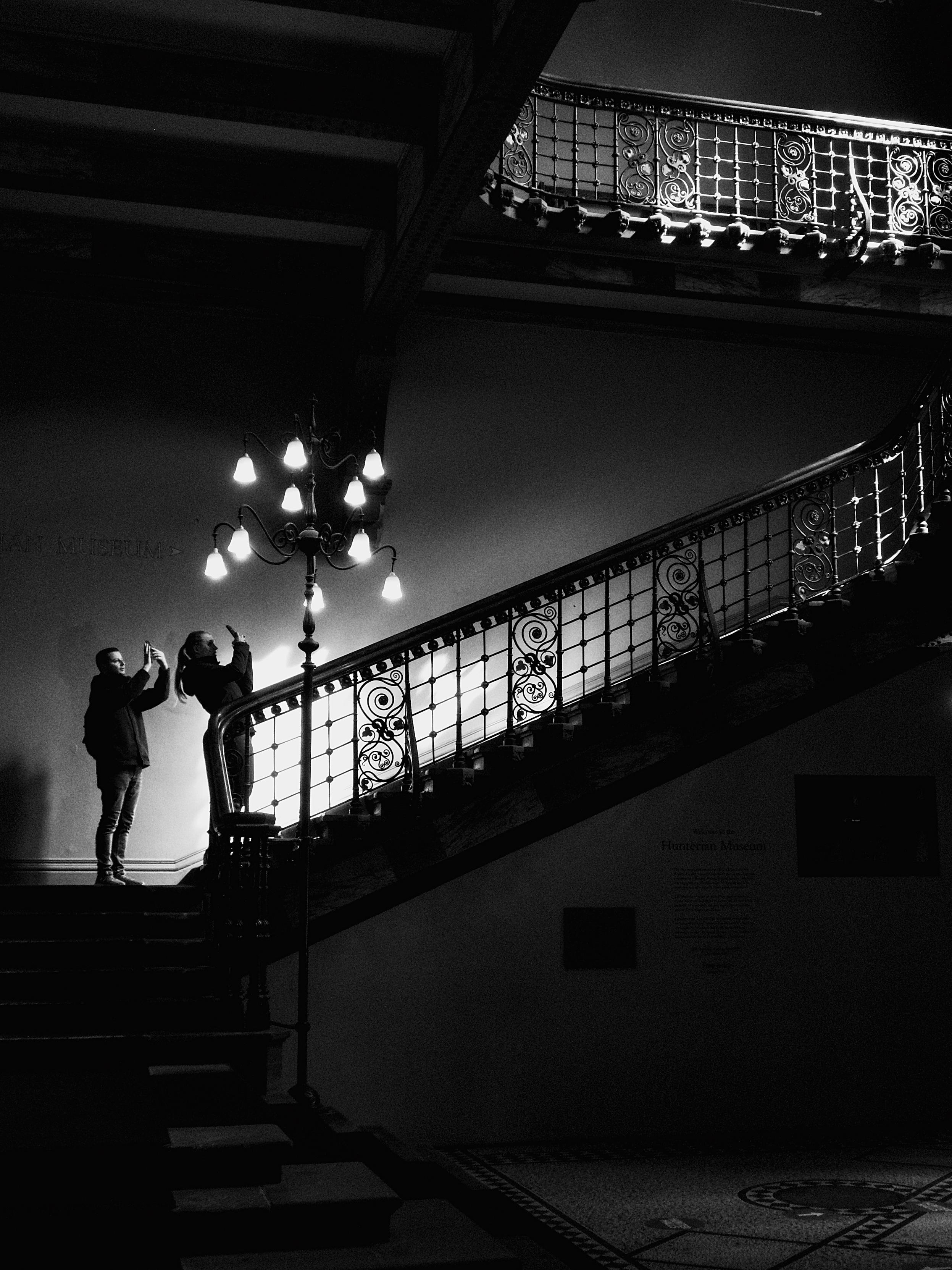 Two people pause to take phone photos on the grand steps of the Hunterian Museum at Glasgow University. Sunlight through a tall, decorative window illuminates the railings on the upper floor, while small ornate lamps at the foot of the first flight cast a soft glow.