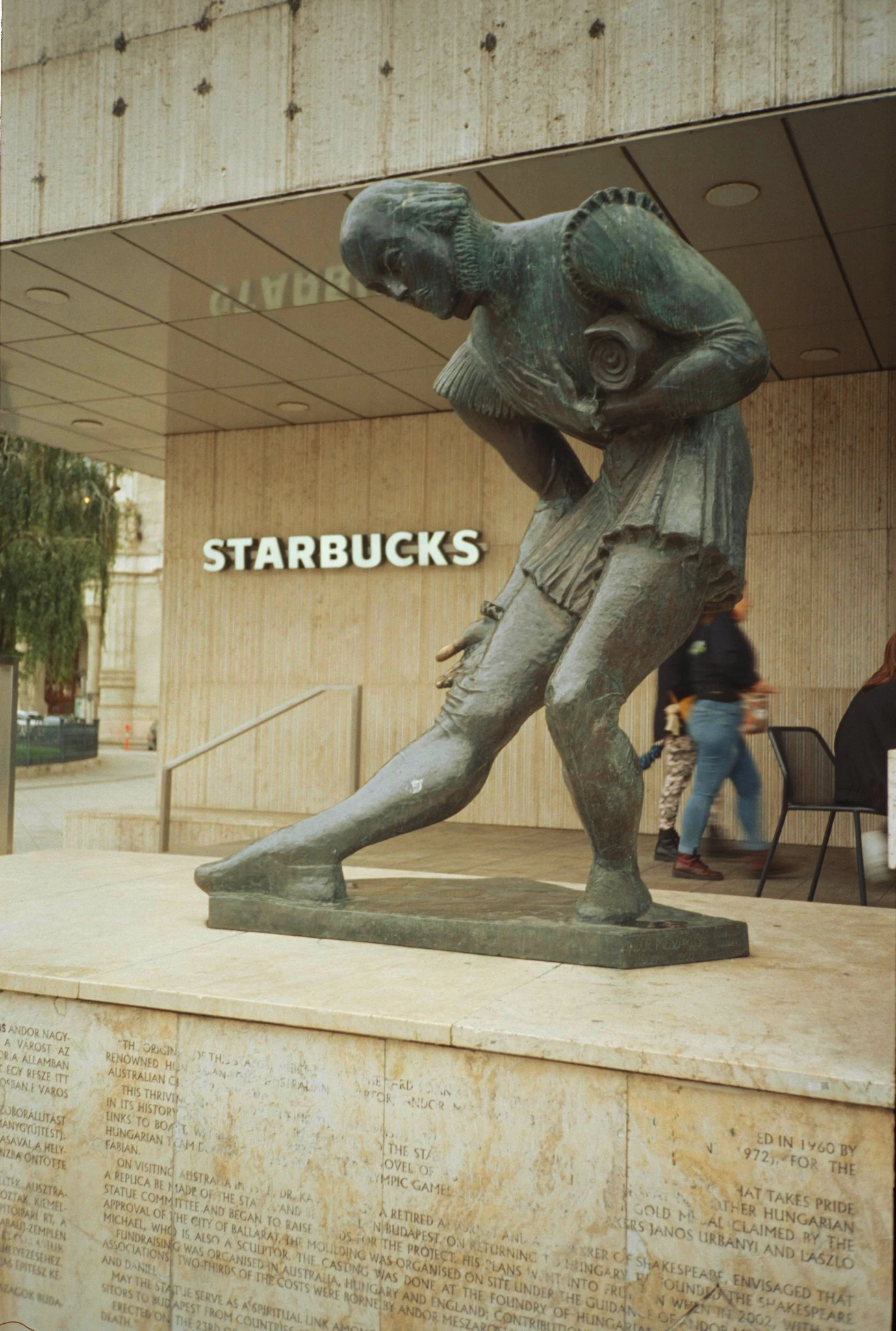 eine bronzestatue zeigt William Shakespeare, der künstler ist Andor Mészáros, die statue ist merkwürdig verrenkt, als hätte er schmerzen im knie. dahinter ein haus und daran große lettern STARBUCKS. die statue steht in budapest Vigadó tér