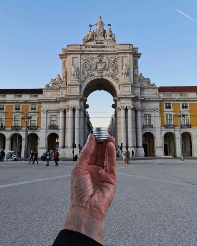 A forced-perspective photo features the historic Rua Augusta Arch (Arco da Rua Augusta) in Lisbon, Portugal, aligned to look like a giant lightbulb base held in a person's hand.