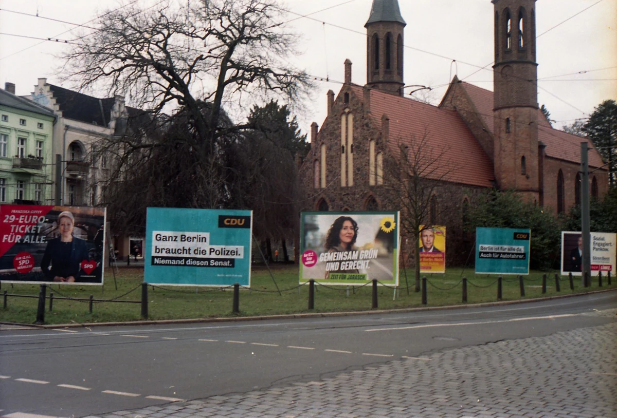 ein etwas unscharfes und unterbelichtetes pfoto einer kirche, davor große wahlplakate von spd, cdu, grüne usw