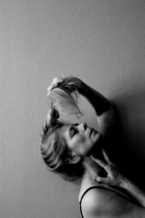 A woman seen in profile leaning her head back with her eyes closed looking quite pleased. She is holding one arm up against her neck and the other hand above her head, holding her twisted hair in her hand. It is a black and white photo. 
