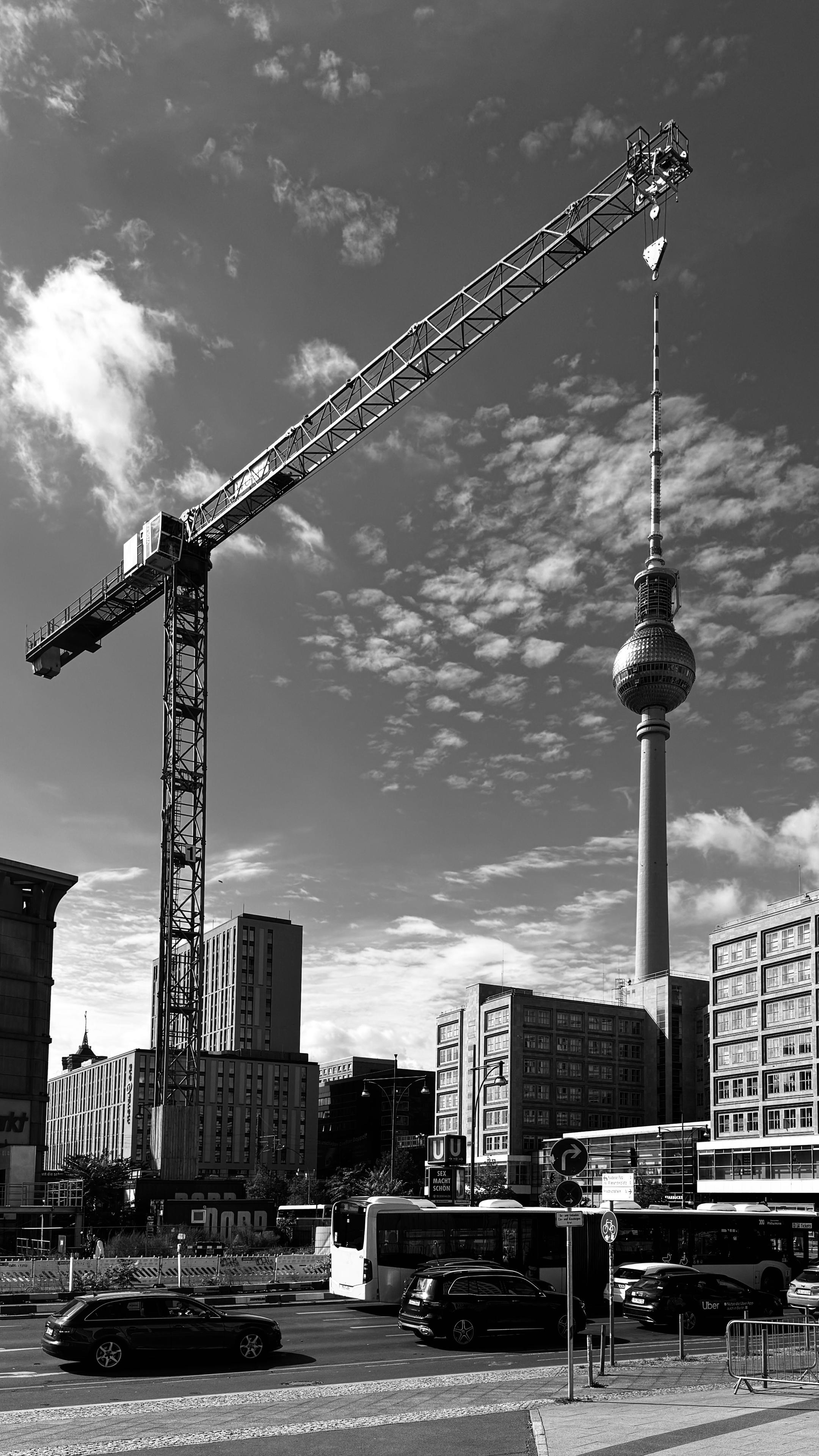 A black and white image featuring a construction crane beside the Berlin TV Tower. Urban buildings and vehicles can be seen below, with a cloudy sky in the background.