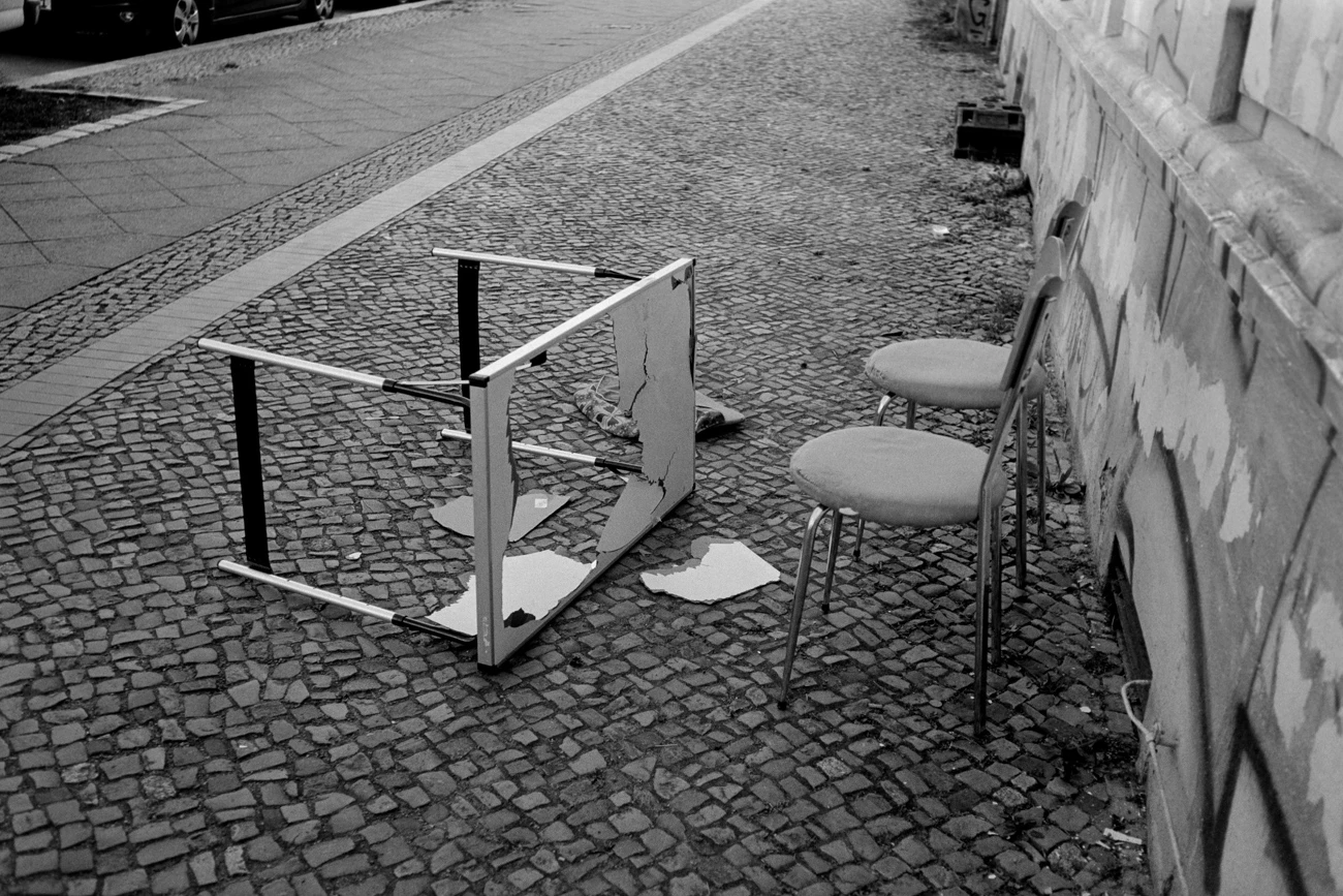 eine schwarzweiße fotografie eines gepflasterten bürgersteigs im urbanen umfeld. rechts deutet sich ein mehrfamilienhaus an. auf dem bürgersteig ohne kontext zwei küchenstühle und ein umgefallener tisch davor. aus der tischplatte sind teile herausgebrochen, das material ist nur als plastik erahnbar.